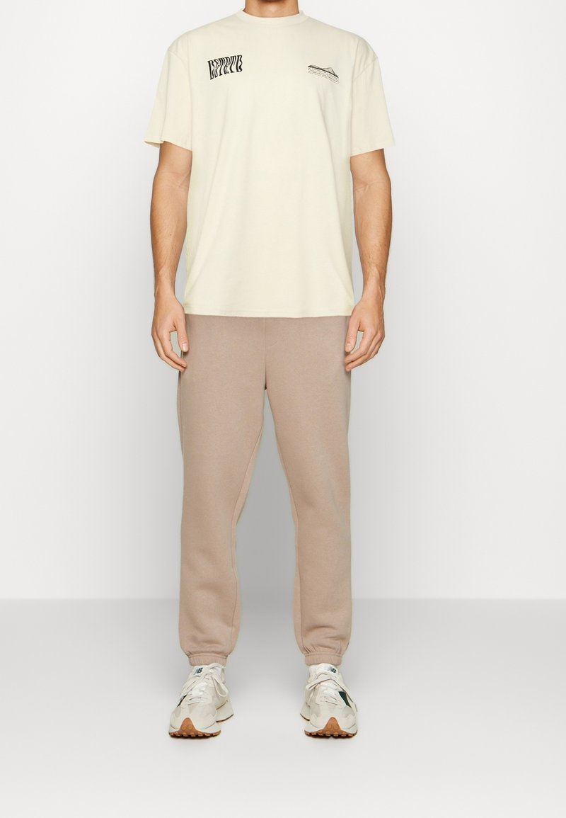 Homme portant un t-shirt graphique crème, un pantalon de jogging beige et des baskets blanches, debout devant un fond blanc uni.