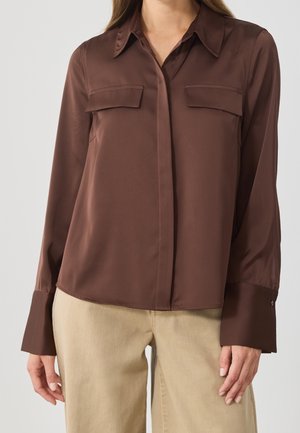 Kobieta ubrana w ciemnobrązową koszulę na guziki z długim rękawem i kieszeniami na piersi oraz beżowe spodnie, stojąca na jednolitym tle.