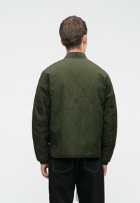 Tamsiai žalia, pakloto tipo bomber tipo striukė su raukšlėtu apykakle ir elastingomis rankovėmis, turinti lygią, vandeniui atsparią tekstūrą.