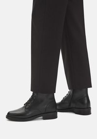 Czarne skórzane botki za kostkę z sznurówkami i bocznymi zamkami, noszone z czarnymi prostymi spodniami na białym tle.