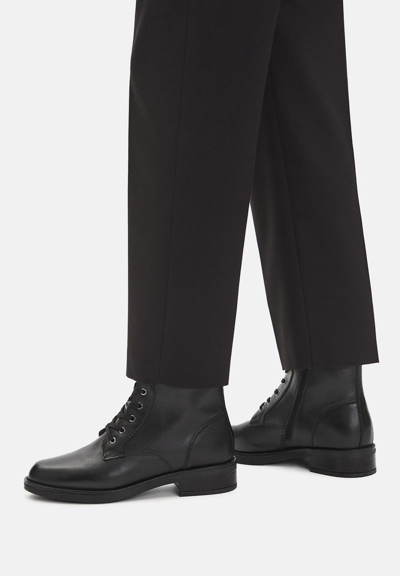 Czarne skórzane botki za kostkę z sznurówkami i bocznymi zamkami, noszone z czarnymi prostymi spodniami na białym tle.