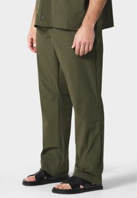 Pantalones de corte relajado en verde oliva, confeccionados en una tela ligera, con perneras rectas y bolsillos laterales, combinados con sandalias negras de tipo slip-on.