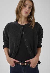 Cárdigan gris oscuro hecho de un material suave, con escote redondo, cierre de botones y puños acanalados, usado sobre una camiseta negra y vaqueros azules.