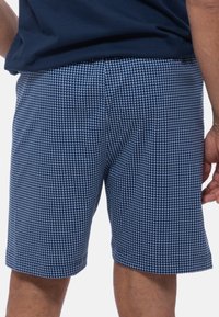 Eine Person trägt ein marineblaues T-Shirt und gemusterte blaue Shorts, steht mit dem Rücken zur Kamera und hat eine Hand auf der Hüfte. Die Shorts verfügen über ein Gittermuster.