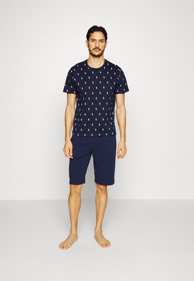 Tricou din bumbac de culoare navy cu un model alb, purtat cu pantaloni scurți asortați. Mâneci scurte, gât rotund și croială lejeră. Poziție desculță.