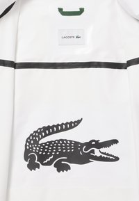 Giacca impermeabile bianca con un distintivo logo di coccodrillo nero. Dettagli a strisce nere sulle spalle. L'etichetta riporta "Lacoste" con un accento verde.