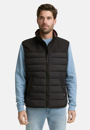 Uomo che indossa un gilet trapuntato nero sopra una felpa azzurra e jeans azzurri, in piedi davanti a uno sfondo bianco uniforme.