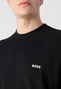 Fekete kötött pulóver, kerek nyakkal, texturált részletekkel és fehér "BOSS" logóval a mellrészen. Sima anyagú felület.