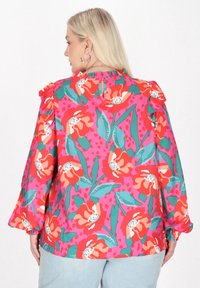 Virágos blúz fodros ujjakkal; élénk rózsaszín alap piros virágokkal és zöld levelekkel; hátul kulcslyuk részlettel és rugalmas mandzsettákkal rendelkezik.