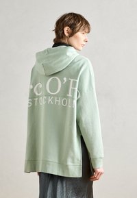 Mintgroene oversized hoodie met witte lettering "STOCKHOLM" en "c O' B" op de achterkant, voorzien van een capuchon met trekkoord en een geribbelde onderkant.