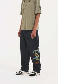 Olive green oversized t-shirt with short sleeves, paired with black jeans featuring a colorful dragon embroidery on the left leg and black sneakers.