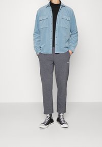 Mann mit hellblauem Cordhemd über schwarzem Oberteil, grauen Jogginghosen und schwarzen High-Top-Sneakern, der mit den Händen in den Taschen steht.