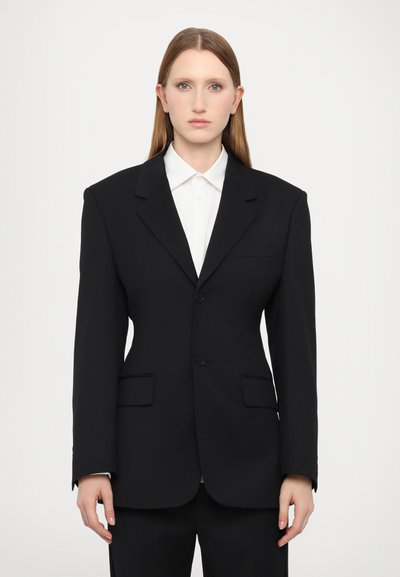 Černý přizpůsobený blazer se strukturovanou siluetou, zapínáním na jeden knoflík, klopovými límci a dvěma předními kapsami. Nosič má na sobě bílou košili.