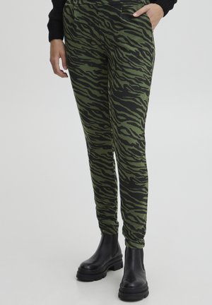Nahaufnahme einer Person, die grüne und schwarze tigerstreifenhose mit seitlichen Taschen und schwarze klobige Stiefeletten vor hellem Hintergrund trägt.