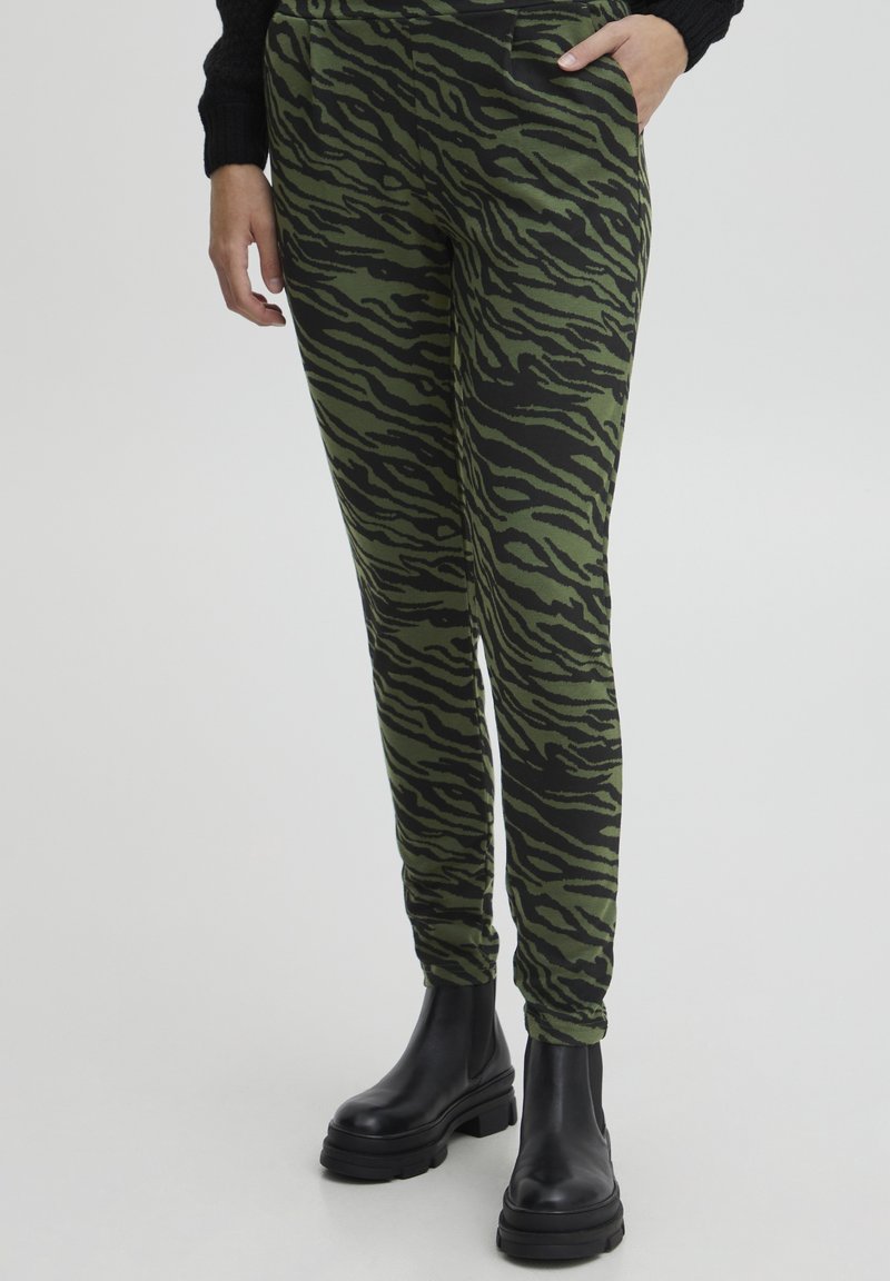 Gros plan d'une personne portant un pantalon rayé tigre vert et noir avec des poches latérales et des bottines noires épaisses sur un fond clair.