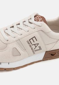 Světlobéžová teniska z použitím semišu a síťoviny, s výrazným logem EA, bílou podrážkou a hnědými detaily na patě a gumové podešvi.