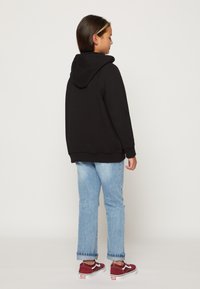 Felpa nera con tasca frontale e cappuccio, abbinata a jeans chiari strappati e sneakers burgundy Vans con dettagli bianchi.