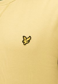 Κίτρινο ύφασμα με ένα μικρό κεντημένο κίτρινο πουλί με μαύρο περίγραμμα ραμμένο κοντά στη ραφή.