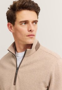 Beige Zip-Hoodie mit einem Fischgrätmuster, das eine braune Akzentuierung am Kragen und eine glatte Textur aufweist.