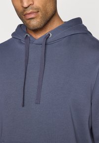 Granatowa bluza z kapturem o miękkiej fakturze, z prążkowanym dołem i sznurkami. Charakteryzuje się luźnym krojem i metalowymi oczkami w kapturze.