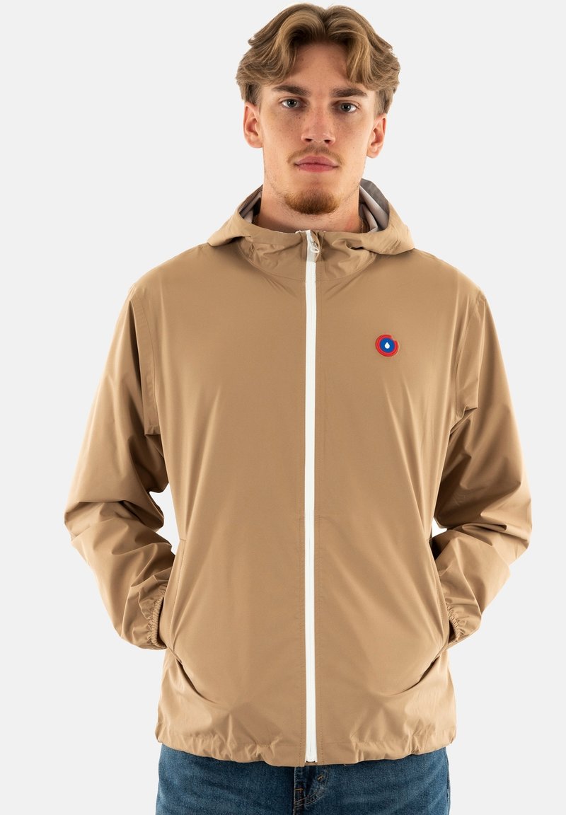 Veste légère beige avec capuche, zip entier et poignets élastiques ; arbore un petit logo circulaire rouge et bleu sur la poitrine.
