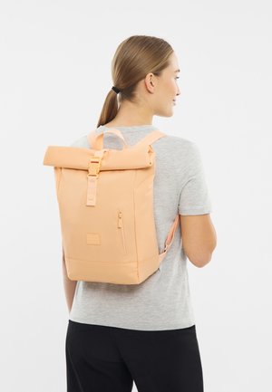 Donna che indossa una maglietta grigia chiara e pantaloni neri, porta sulla schiena uno zaino roll-top color pesca con una tasca con zip frontale.
