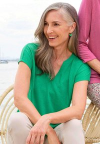 Groen shirt met korte mouwen en V-hals, gemaakt van zacht materiaal, gecombineerd met lichtbeige broek. Verdere details omvatten oorbellen en een subtiele gestructureerde achtergrond.