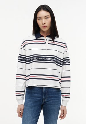 Kobieta nosząca białą koszulkę rugby z długim rękawem z granatowymi i czerwonymi paskami, tekstem „tommy jeans” i ciemnoniebieskim kołnierzem, zestawioną z niebieskimi dżinsami.