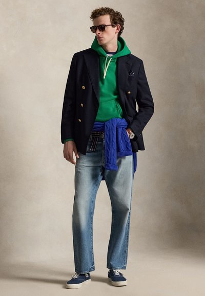 Homme portant un sweat à capuche vert sous un blazer noir, jean bleu, pull bleu noué à la taille, lunettes de soleil et baskets bleu marine, posant devant un fond neutre.