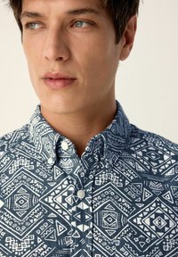 Blau gemusterte Hemd mit Knopfleiste, das abstrakte weiße geometrische Designs zeigt, kurzer Kragen und leichte Stofftextur. Nahaufnahme von Hals- und Brustbereich.