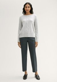 Grauer Kabelstrickpullover mit weitem Ausschnitt, kombiniert mit dunklen Karohosen. Das Model trägt schwarze Slipper. Einfaches Design, weicher Stoff.