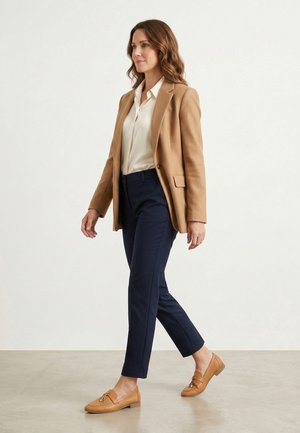 Kvinde går indendørs iført en beige blazer, hvid bluse, marineblå bukser og beige loafers mod en ensartet lys baggrund.