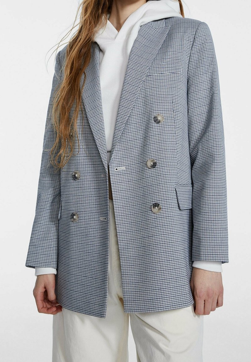 Femme portant un blazer gris à carreaux croisé par-dessus un sweat à capuche blanc et un pantalon couleur crème, debout devant un fond uni.