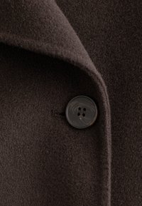 Brauner Wollmantel mit einer weichen, strukturierten Oberfläche. Verfügt über einen klassischen Revers und einen schwarzen Knopf mit vier Löchern, was funktionale Details hinzufügt.