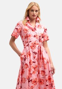 Rochie florală roz cu mâneci scurte, închisă cu nasturi în față, cu un cordon la talie și buzunare. Prezintă flori mari de culoare portocalie și roșie pe un fundal deschis.