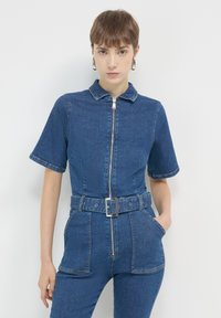 Salopette en denim bleu foncé avec fermeture éclair à l'avant, manches courtes, taille ceinturée et deux poches avant. Présente un col et un tissu texturé.