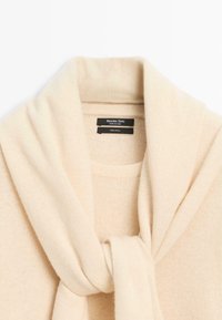 Kremowy sweter wełniany z dużym, owiniętym kołnierzem o fakturowanym wykończeniu. Metki wskazują, że materiał to 100% wełna.