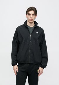 Μαύρο μπουφάν με φερμουάρ, με ψηλό γιακά, διακριτικές ραφές, λογότυπο μπροστά και ελαστικά μανίκια, φορεμένο πάνω από σκούρο πουκάμισο και παντελόνι.