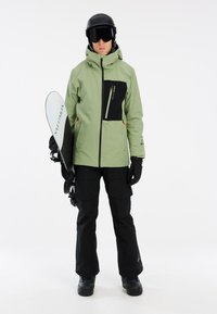 Giacca impermeabile verde con accenti neri, zip frontale e tasche. Pantaloni neri e guanti. Persona che tiene uno snowboard. Casco nero e occhiali protettivi.