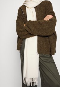 Person mit verschränkten Armen, die einen braunen Reißverschluss-Pullover, olivgrüne Hose und einen langen cremefarbenen Schal mit fransigen Enden trägt.