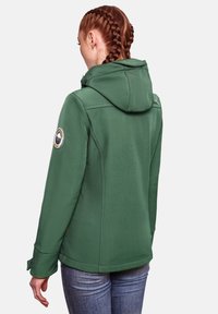 Žalia gobtuvė su lietuviu audiniu, turi logotipo atvartą ant rankovės ir pritaikytas siūles, derinama su mėlynomis džinsomis.