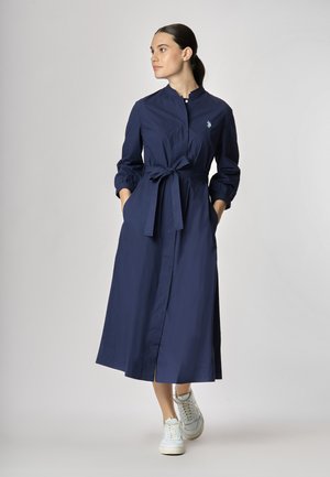 Abito lungo blu navy con maniche lunghe, colletto alla mandarino e vita con cintura. Dotato di tasche laterali e uno spacco sul fondo. Indossato con sneakers bianche.