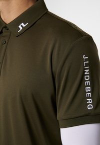 Olivi vihreä polo paita, jossa on kaulus ja kolme nappia. Vasemmassa hihassa on valkoisella brodeerattu "J.LINDEBERG".