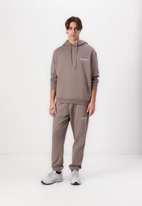 Brun hættetrøje med snoretræk og frontlomme, parret med matchende joggers. Begge har logo detaljer. Model iført grå træningssko.