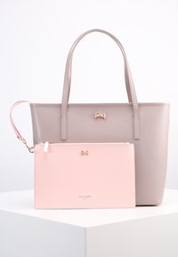 Jasnoszara torba tote o fakturze z teksturą, ozdobiona złotą kokardą, w komplecie z miękkim różowym etui, które również ma złotą kokardę i logo.