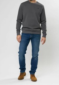 Sudadera gris con cuello redondo y puños de canalé, combinada con jeans azules y botas de ante marrón. Pequeño logotipo en el pecho.
