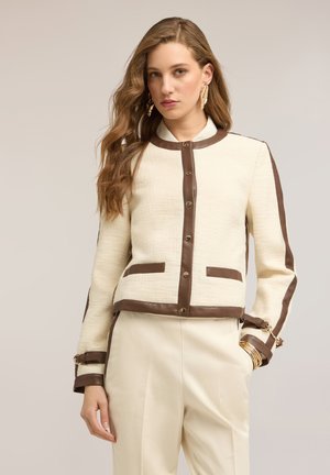 Kobieta z długimi, falowanymi włosami, ubrana w kremową kurtkę z brązowymi wykończeniami, złote kolczyki w kształcie liści, kremowe spodnie i złote bransoletki, pozująca z jedną ręką w kieszeni.