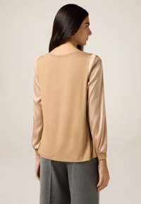 Top beige con maniche in raso, scollatura rotonda, maniche lunghe con polsini arricciati e vestibilità comoda. Abbinato a pantaloni grigi a gamba larga.