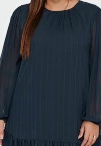 Marinblå klänning med långa genomskinliga ärmar, en samlad halsringning och vertikal randstruktur. Uppsvept nederkant med lager av volangdetaljer.