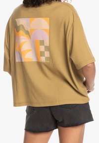 Avslappnad passform, ribbad senapsgul t-shirt med en färgglad geometrisk design på baksidan. I kombination med svarta denimshorts, vilket ger en avslappnad stil.